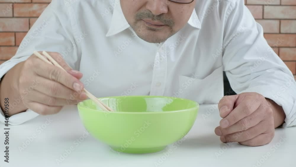 Hungry Thai man in white shirt uses chopsticks to eat hot instant noodles in green bowl in lunch breaks, quick, tasty, and cheap. Traditional Asian fast food meal of Japanese and Chinese lifestyle. Hungry Thai man in white shirt uses chopsticks to eat hot instant noodles in green bowl in lunch breaks, quick, tasty, and cheap. Traditional Asian fast food meal of Japanese and Chinese lifestyle.