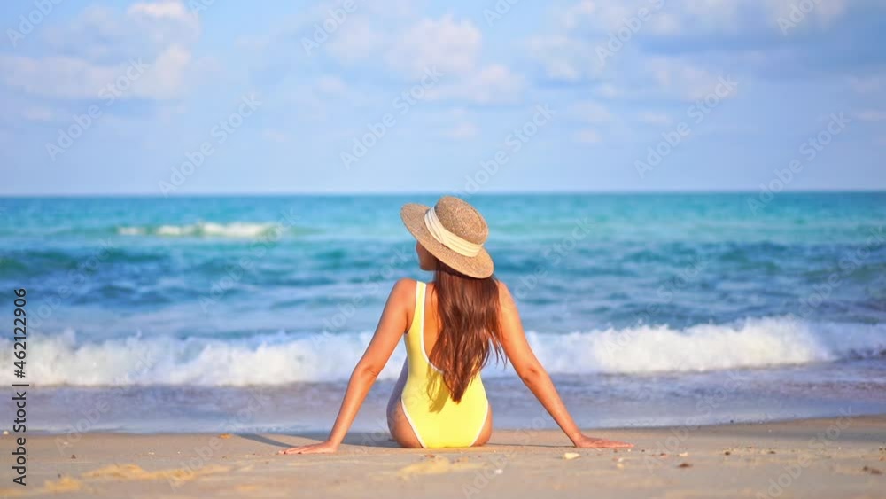 Back view of woman sitting on sandy beach with yellow swimsuit and hat. Slow-motion