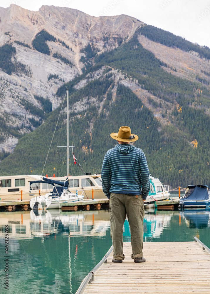A man with a hat standing on a pier by a beautiful lake and mountains in the background.