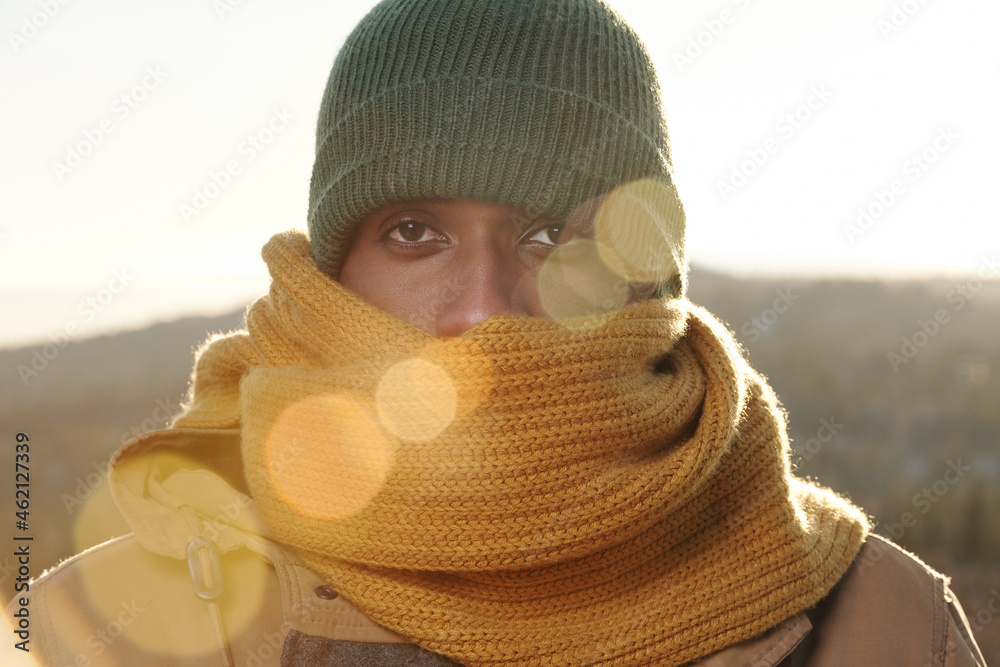 Frozen young man of African ethnicity in knitted scarf and cap looking at camera - obrazy, fototapety, plakaty 