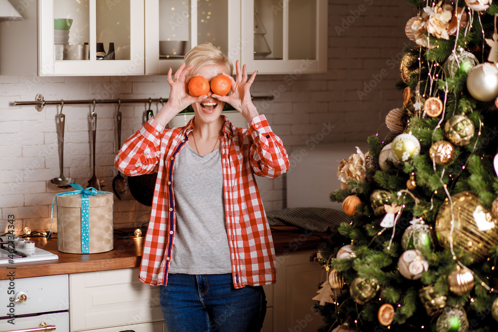 A beautiful girl in a red plaid shirt and blue jeans stands near the Christmas tree and covers her eyes with two tangerines. Happy and joyful emotions. Christmas celebration.