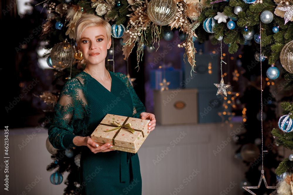 Beautiful girl in a green dress with a gift in hands on Christmas and New Year's Eve. Portrait smilling girl near Xmas tree. Christmas celebration. Girl decorate living room for Christmas.