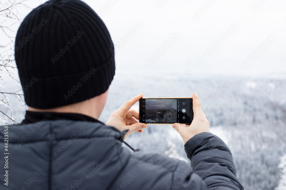 Obraz premium Close-up of a man photographing a winter landscape on the phone. Mobile photography. Winter tourism and hiking in the forest. Copy space