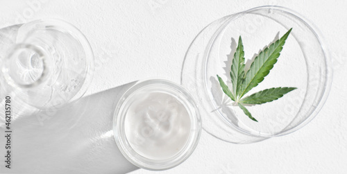 CBD cream in glass jar and hemp leaves on petri dish on white table. Marijuana as cosmetic ingredient. using hemp leaves extract in cosmetic for beautiful skin. top view. banner.