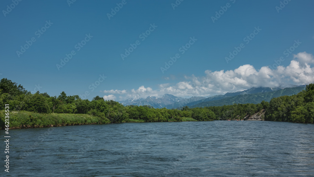 The blue river flows peacefully. In the distance, inflatable boats with people. On the banks of lush vegetation, wildflowers. Picturesque mountains on the background of azure sky and clouds. Kamchatka