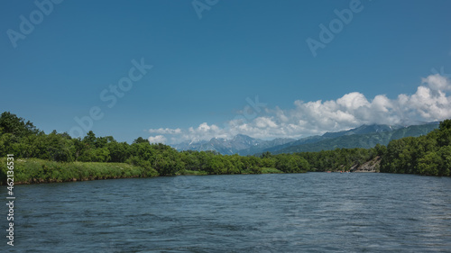 The blue river flows peacefully. In the distance, inflatable boats with people. On the banks of lush vegetation, wildflowers. Picturesque mountains on the background of azure sky and clouds. Kamchatka