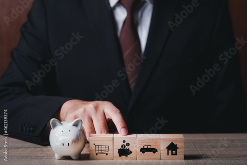 businessman holding finger point at piggy bank wooden block goal target plan for saving money with shopping, car and home. copy space for text.