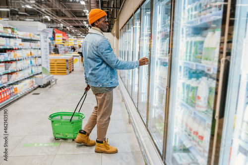 African-American guy with green plastic basket walks looking at shelves with different goods in modern supermarket department