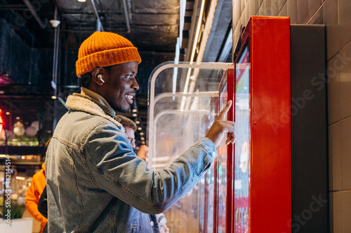 Positive African-American guy in orange knitted hat with wireless earphones orders food through self-service kiosk in cafe photo