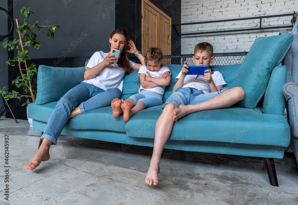 Adult woman and two children are sitting on blue sofa. Mom and son use phone and do not pay attention to girl sitting next to them. Adult woman and two children are sitting on blue sofa. Mom and son use phone and do not pay attention to girl sitting next to them.