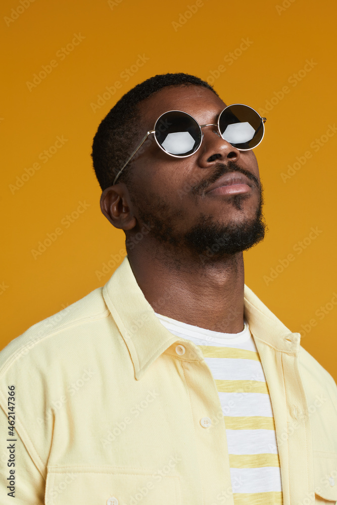 Fototapeta premium Vertical portrait of handsome African-American man wearing sunglasses while posing against yellow background in studio