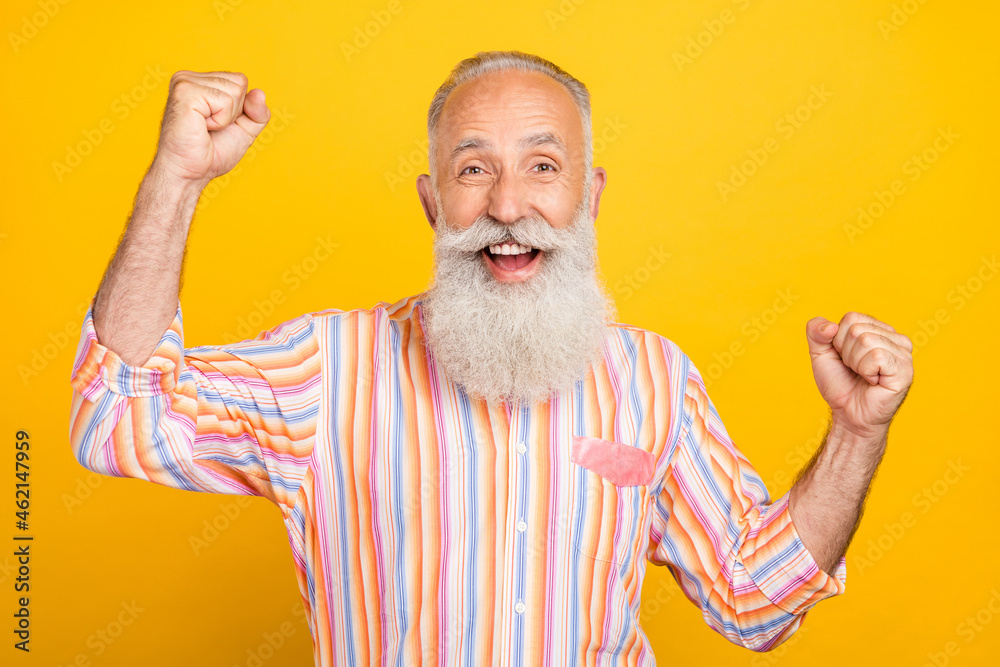 Photo of lucky funny age gentleman wear striped shirt smiling rising fists isolated yellow color background Photo of lucky funny age gentleman wear striped shirt smiling rising fists isolated yellow color background