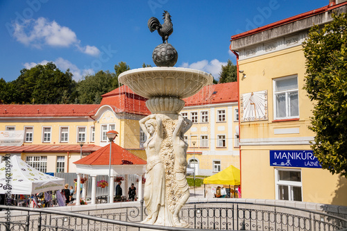 Lazne Libverda, North Bohemia, Czech Republic, 04 September 2021: fountain with women and rooster near red and white colonnade at sunny summer day, historical spa, city fair selling food, souvenirs