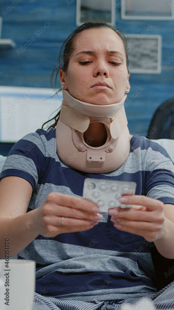 Sick woman with cervical neck foam reading label of medication pills against disease. Adult wearing medical collar for injured muscle while looking at tablet with capsules for treatment