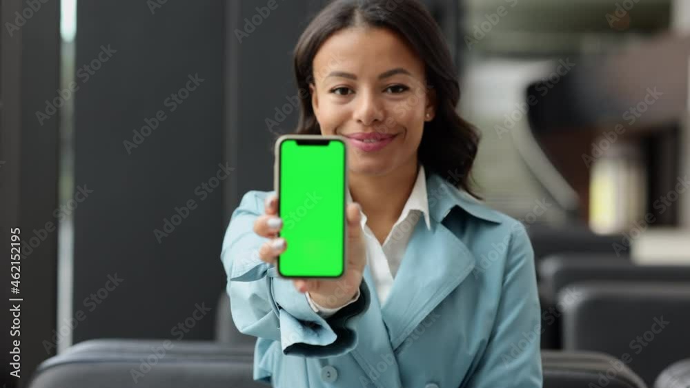 Young businesswoman expecting flight and showing smartphone while sitting in airport room spbi. Close front view of American African woman uses phone and sits at interior, then shows screen and looks Young businesswoman expecting flight and showing smartphone while sitting in airport room spbi. Close front view of American African woman uses phone and sits at interior, then shows screen and looks