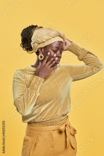 Vertical portrait of trendy African-American woman wearing head wrap and ethnic jewelry while dancing against yellow background in studio © Seventyfour