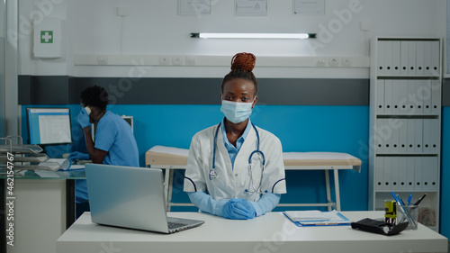 Close up of woman working as doctor in medical cabinet at facility. Portrait of young medic wearing face mask and sitting at desk with laptop  documents  files  and checkup papers