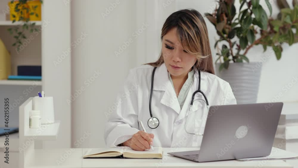 Female doctor working with laptop and writing while sitting at table in clinic office spbi. Front view of young Asian female types on keyboard and looks at computer screen, writes in notebook and sits Female doctor working with laptop and writing while sitting at table in clinic office spbi. Front view of young Asian female types on keyboard and looks at computer screen, writes in notebook and sits