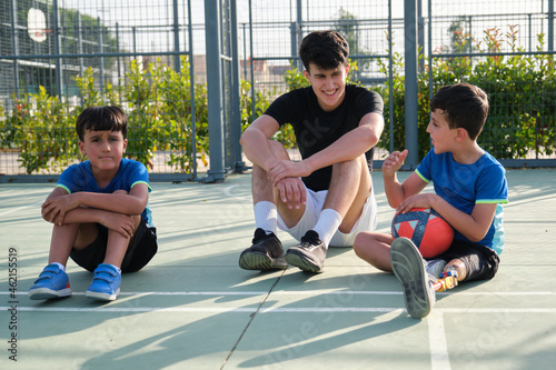 A coach sit on the football court floor resting and talking with two students.