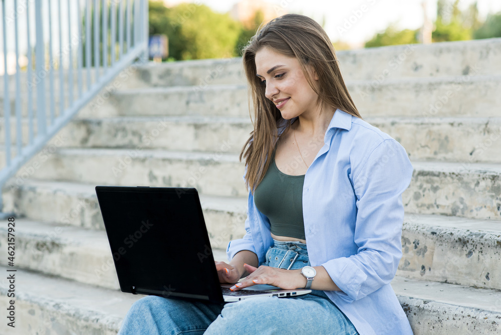 A happy and attractive Caucasian girl is sitting with a laptop on the steps. Distance working. Freelance concept A happy and attractive Caucasian girl is sitting with a laptop on the steps. Distance working. Freelance concept