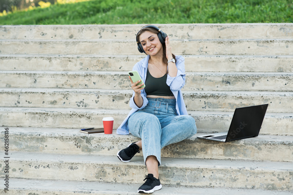 Obraz premium Happy and attractive caucasian girl sitting with a laptop on the steps and holding a phone, listening to music on headphones