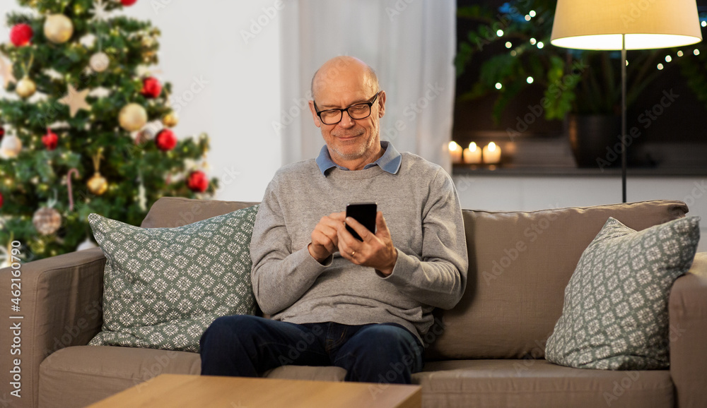 technology, people and winter holidays concept - happy smiling bald senior man texting on smartphone at home in evening over christmas tree on background