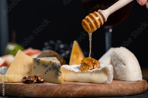 Honey is poured from a special spoon onto Camembert with walnuts. Various types of cheese lie around the delicacy. Close-up, dark background.