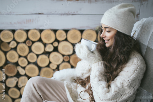Young beautiful woman wearing knitted hat and white sweater drinking tea sitting on armchair on porch of cozy house.