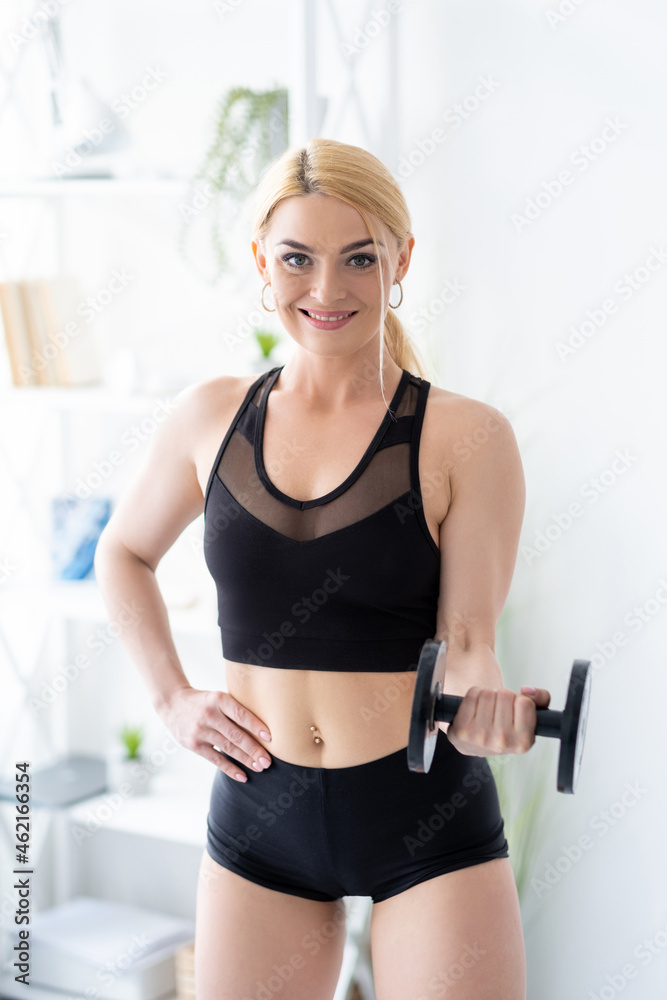 Bodybuilding gym. Female trainer. Home fitness. Strong body. Happy athletic woman in black sportswear doing dumbbells workout for hand biceps in light room interior.