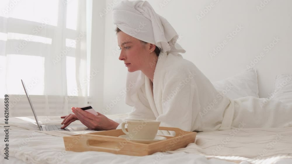 Woman buying online and using laptop while lying on bed in home room indoors spbi. Young female holds card in hand and looks at computer screen, makes purchases in online store and lies in white Woman buying online and using laptop while lying on bed in home room indoors spbi. Young female holds card in hand and looks at computer screen, makes purchases in online store and lies in white