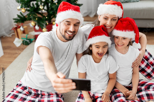 family  winter holidays and christmas concept - happy mother  father and two daughters in santa hats at home taking selfie with smartphone