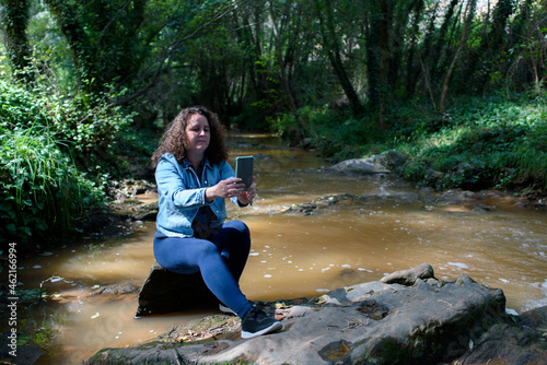 Woman sitting on a rock next to a stream running through a gallery forest, while taking a selfie with her smartphone.