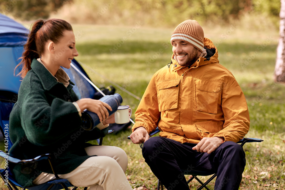 Fototapeta premium camping, tourism and travel concept - happy couple with thermos pouring tea to cup at tent camp