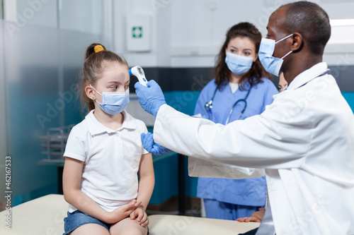 African american pediatrician doctor with face mask against coronavirus measuring girl temperature using medical infrared thermometer. Therapist doctor working in hospital office. Health care service