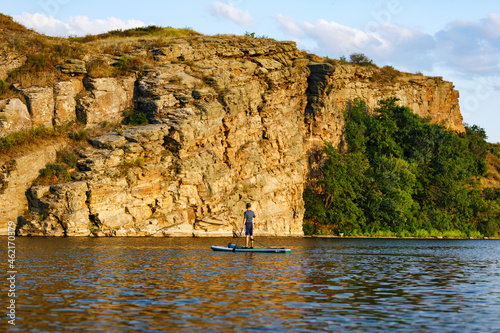 A man on a SUP on the river in summer at sunset against the backdrop of a rock