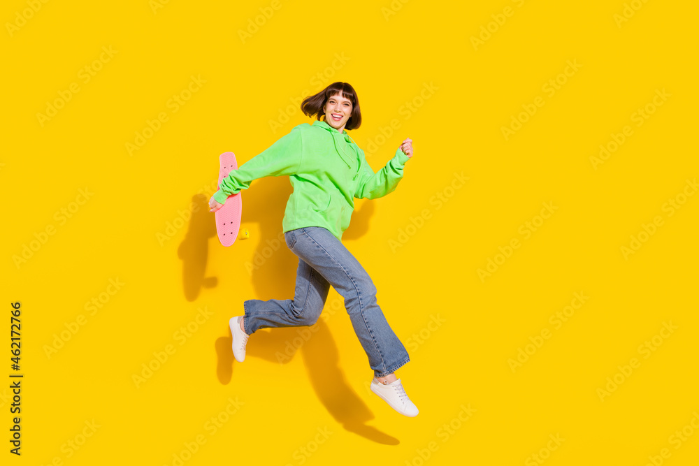 Full length body size view of pretty cheerful girl jumping carrying longboard isolated over bright yellow color background Full length body size view of pretty cheerful girl jumping carrying longboard isolated over bright yellow color background