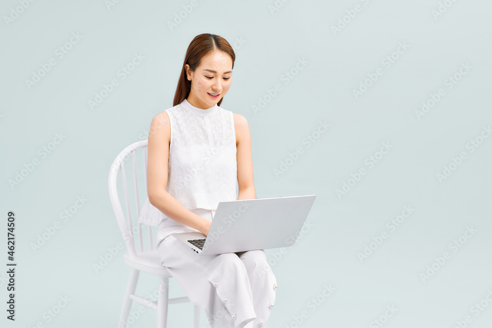 Full length of a happy young woman casually dressed sitting on a chair isolated over white background, working on laptop computer Full length of a happy young woman casually dressed sitting on a chair isolated over white background, working on laptop computer