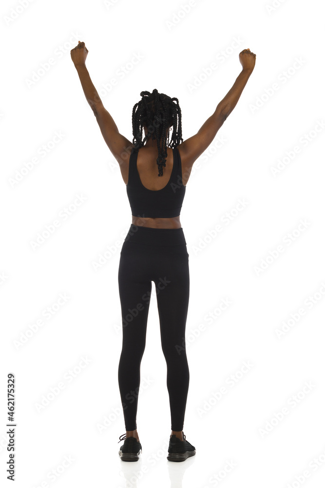 Confident black woman in sports clothes is standing with arms raised and cheering. Full length, rear view.