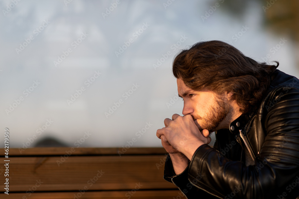 Fototapeta premium Pensive serious young bearded man wear black leather jacket sitting and thinking outdoors