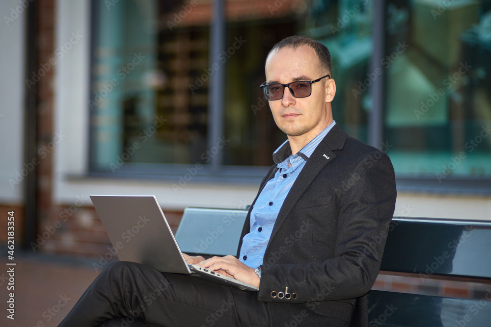Young businessman with glasses and a laptop sitting on a bench outdoor Young businessman with glasses and a laptop sitting on a bench outdoor
