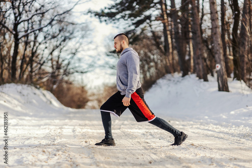 Fit sportsman doing stretching and warmup exercises while standing on snowy path in nature at winter. Healthy habits, winter sport, outdoor fitness