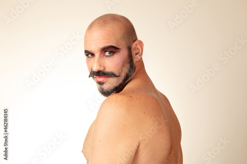 Portrait of bald man without shirt and makeup looks at camera with bare back.  Image in low angle. Side view of caucasian drag man with beard.