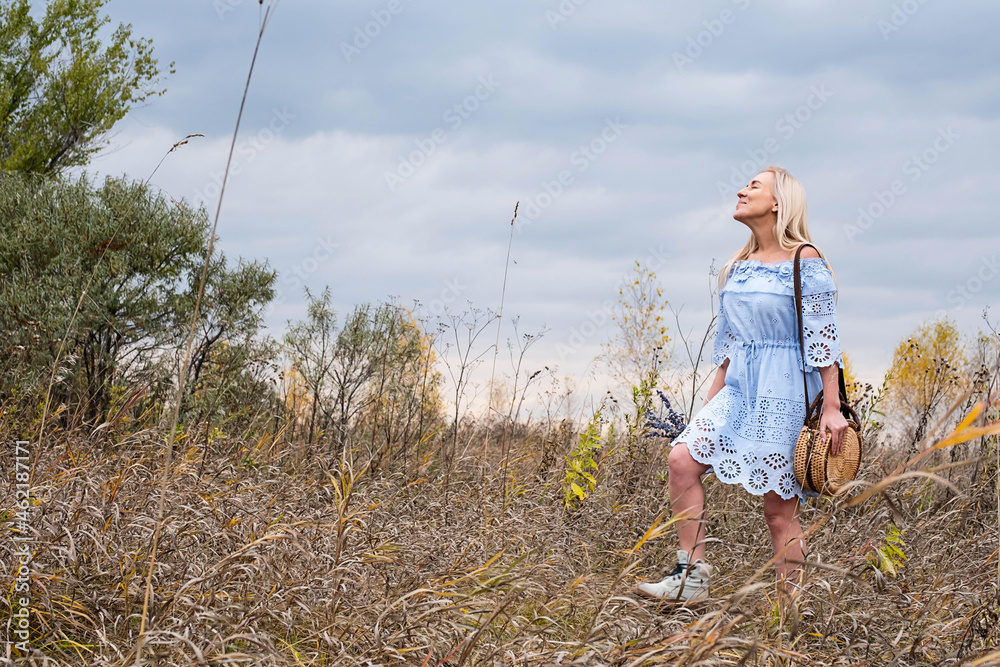 Beautiful blondie woman with trendy nude rattan round bag walking outdoors. Village landscaped, fall season. Beautiful blondie woman with trendy nude rattan round bag walking outdoors. Village landscaped, fall season.