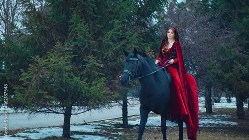 Medieval woman princess in red dress sits astride black steed horse. Girl rider in vintage cloak cape train flies in wind motion. Background green trees spruce forest, spring winter nature melted snow
