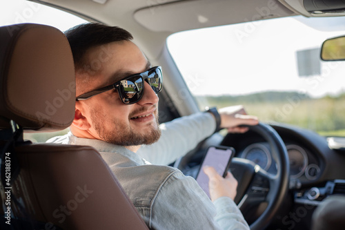 Portrait of young male driver using his mobile phone while driving the car and smiling. Man driver using smart phone in car. © Julija