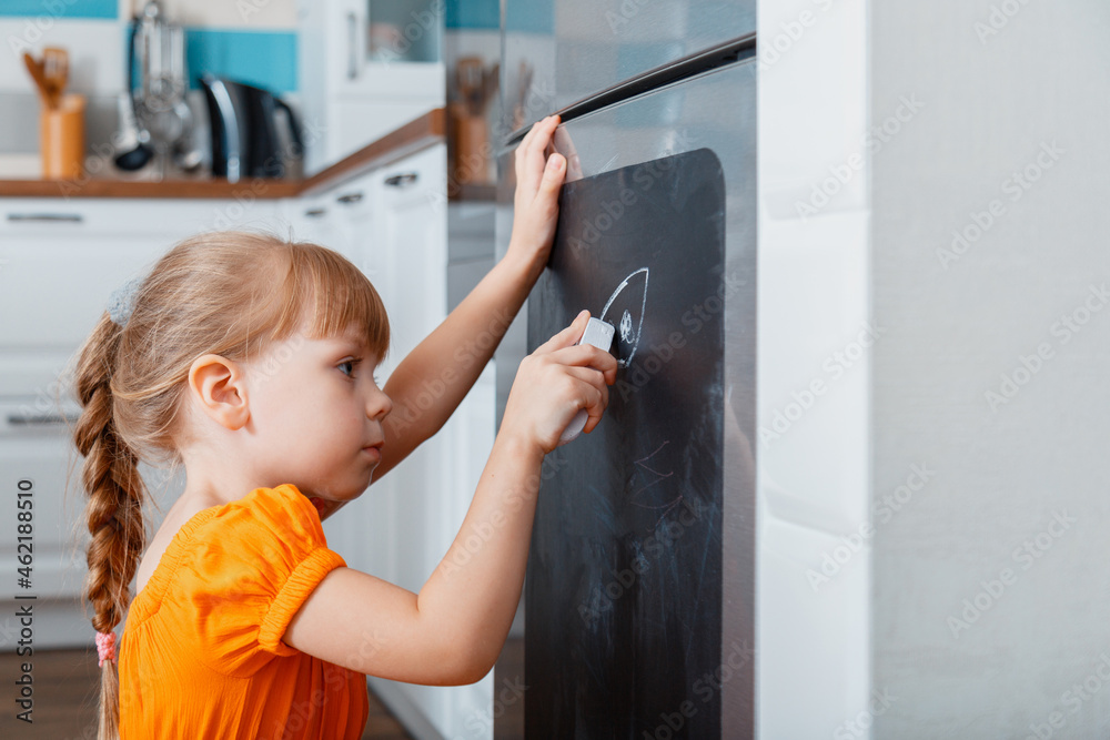 Little caucasian focused child girl draw with chalk on chalk board on refrigerator in kitchen at cozy home interior. Little caucasian focused child girl draw with chalk on chalk board on refrigerator in kitchen at cozy home interior.
