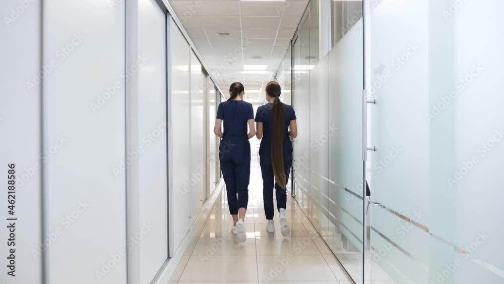 Two female doctors in medical blue suits with long beautiful hair walk in the corridor of a modern clinic discussing the medical history of a patient with heart failure work issues. Two female doctors in medical blue suits with long beautiful hair walk in the corridor of a modern clinic discussing the medical history of a patient with heart failure work issues.