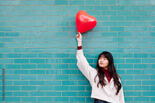 Young woman looking away while holding red heart shape balloon in front of brick wall photo