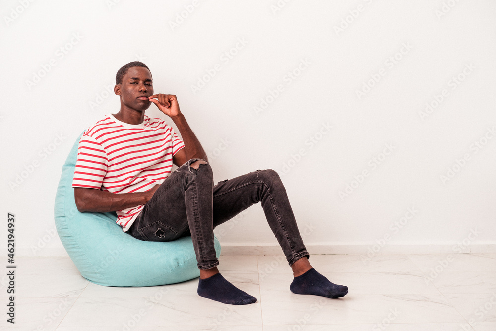 Fototapeta premium Young African American man sitting on a puff isolated on white background with fingers on lips keeping a secret.
