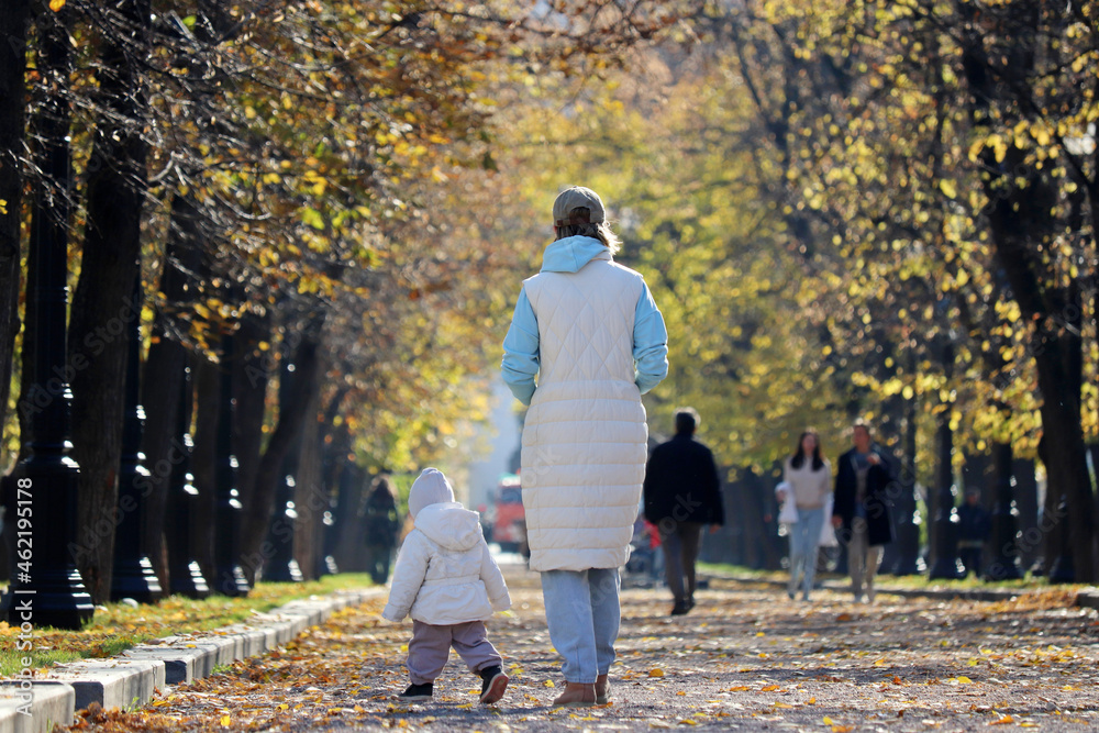 Fototapeta premium Mother and kid are walking down in autumn park. Young slender woman and running child, concept of motherhood, single mother or babysitter